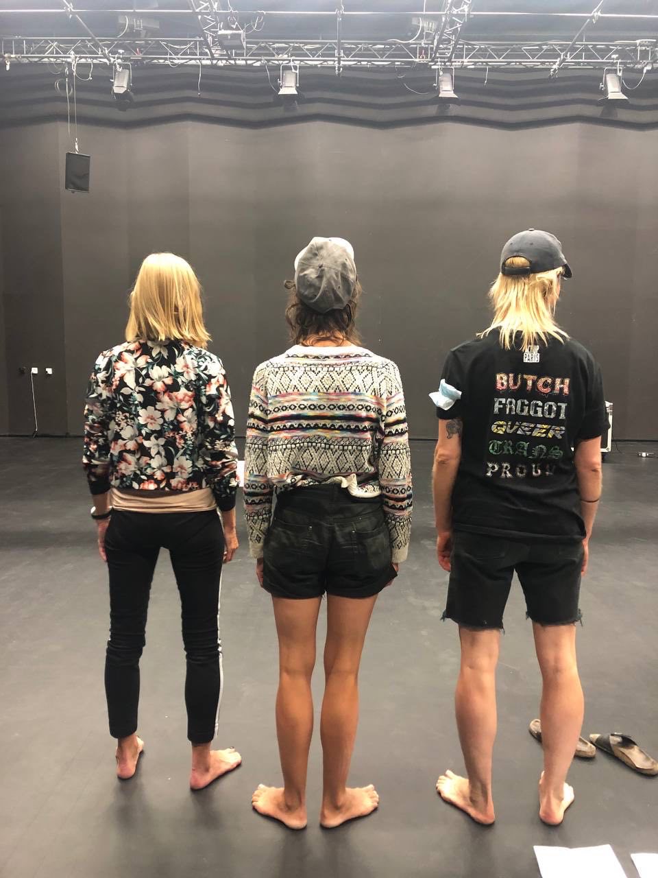 Photo de répétition du spectacle ALKOHOL : trois interprètes vus de dos sur une scène de théâtre, sous les projecteurs et la structure technique de la salle